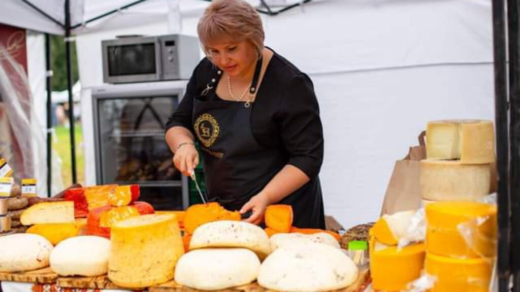 Fromagère ukrainienne en tablier noir découpant du fromage artisanal sur un étal de marché présentant une variété de fromages